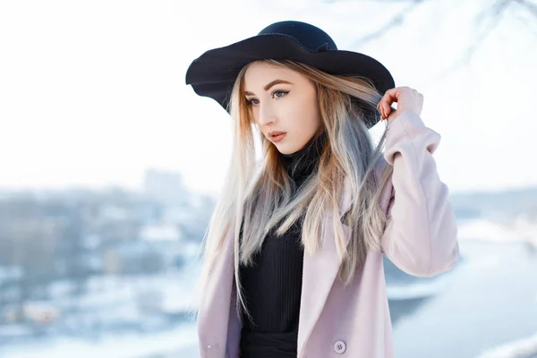 Charming elegant young woman in a vintage hat in retro style in a pink coat in a knitted dress stands on a warm winter day against the background of the river. Sensual girl enjoys nature.