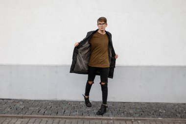 Genç yakışıklı hipster adam sökük siyah kot pantolon siyah spor ayakkabı beyaz duvara yakın poz bir t-shirt moda uzun sonbahar ceket şık bardaklarda. Kentsel Amerikalı adam.