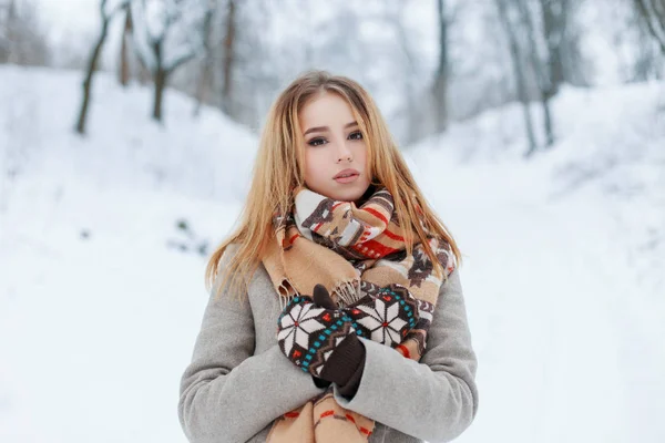 冬の寒い日に雪の降る公園でパターンの残りとスタイリッシュなベージュのマフラーとニットのヴィンテージ ミトンでおしゃれなグレーのコートで美しい魅力的な若い女性 魅力的な少女金髪 — ストック写真
