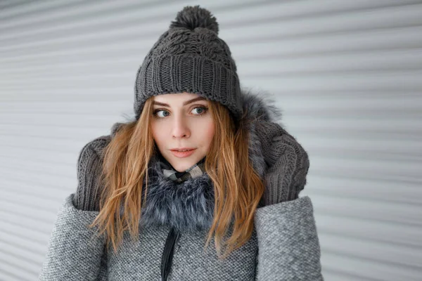 美丽可爱的年轻女子与蓝色的眼睛在针织帽子在灰色的冬衣与毛皮在老式温暖的手套附近的白色金属墙 美丽的女孩在散步 — 图库照片
