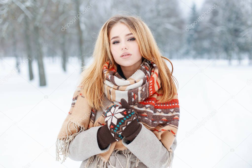 Young woman with beautiful eyes with pale pink lips with a vintage stylish scarf in a warm coat with knitted mittens posing on the background of winter trees in the park. Fashionable modern girl.