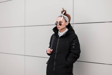 Modern Genç hipster kadın deri sırt çantası ile şık bir bandana şık bir ceket içinde yuvarlak güneş gözlüğü beyaz bir duvar bir sıcak kış günü duruyor. Şık Amerikan Kız tatilde