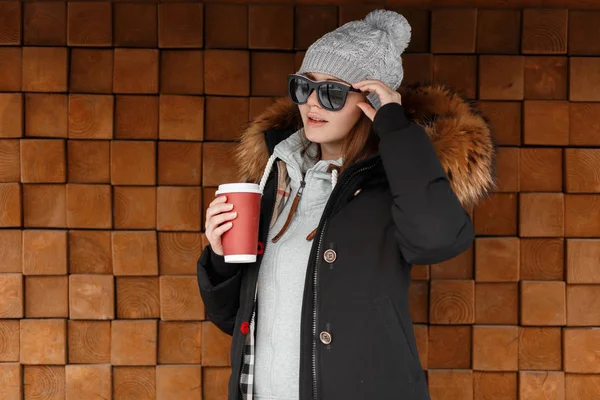 迷人的时尚年轻女子嬉皮士戴着太阳镜 戴着针织帽子 穿着带皮罩的冬季夹克站在户外 手里拿着一个红色的杯子和美味的热咖啡 美丽的美国女孩 — 图库照片