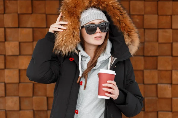 年轻的嬉皮士妇女在一个针织灰色帽子 在一个时尚的冬季夹克与毛皮在温暖的蓝色运动衫拿着一杯咖啡附近的木墙 冬季服装 — 图库照片