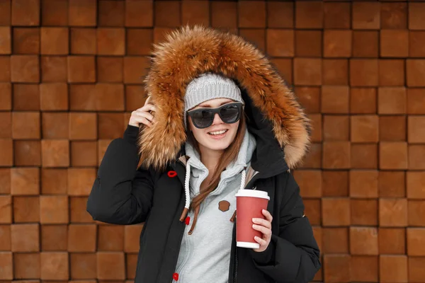 穿着复古针织帽子 穿着皮草的冬季夹克的快乐年轻嬉皮士女子站在木墙附近 手里拿着一个红色的杯子和热咖啡 愉快的快乐的女孩 — 图库照片