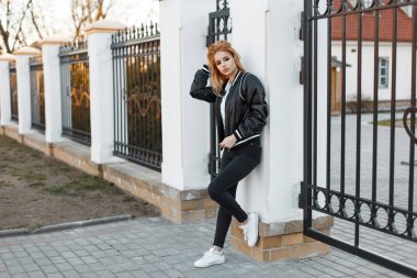 Vintage bir metal çit poz spor ayakkabı içinde beyaz bir polo gömlek modaya uygun kot pantolon Siyah bahar cekette şık genç kadın. Amerikan şık kız. moda