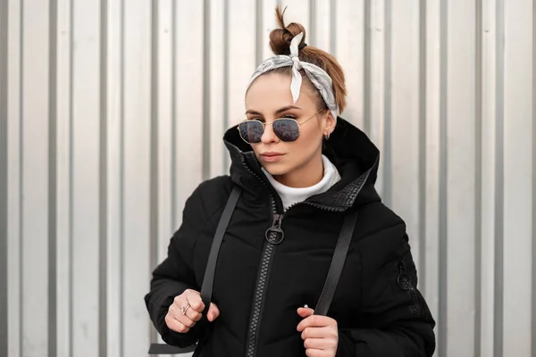 Young attractive hipster woman with stylish hairstyle with a bandana in black glasses in a long black coat with a backpack on her shoulders posing near a metal wall. Stylish girl.