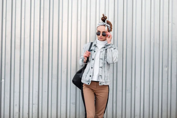 黒のバックパックとスタイリッシュなパンツのデニム ジャケットにニットのヴィンテージ セーターでおしゃれなバンダナでスタイリッシュな髪型でかわいい若い女性流行に敏感な金属製の壁のそばに立ちます 散歩のための女の子 — ストック写真