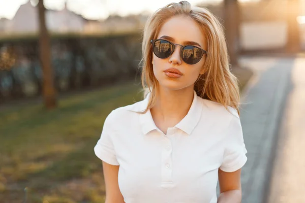 时尚美丽的年轻嬉皮士女子穿着时髦的太阳镜 身穿白色马球 站在街头 背景是夏日的日落 可爱的时尚女孩金发 — 图库照片