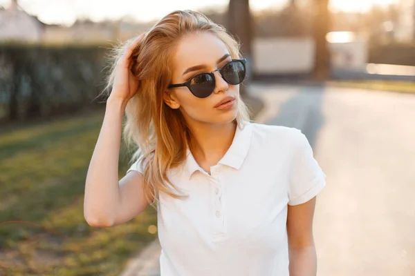 现代时尚的年轻嬉皮士女子在时尚的白色马球 T恤在黑色太阳镜站在街上 并在阳光的背景下摆姿势 美国美丽的女孩 — 图库照片