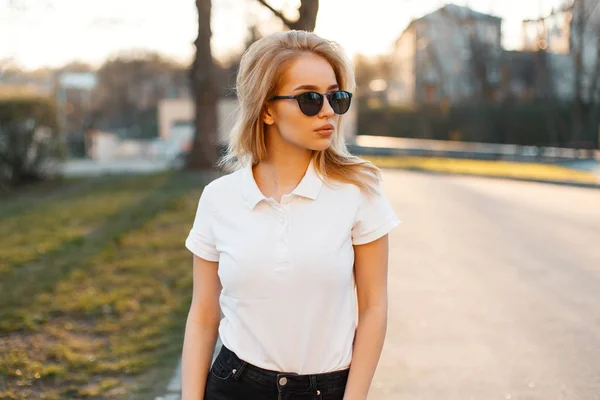 Young attractive hipster woman in stylish sunglasses in a white polo t-shirt in black jeans walking in downtown at sunset. Cute stylish girl. Summer evening.