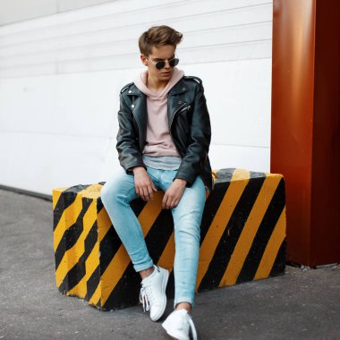 Bir şeritli siyah-sarı betonarme döşeme açık havada beyaz metal kapıları yanında oturan bir saç modeli ile moda giysiler içinde siyah güneş gözlüğü şık yakışıklı genç adam. Amerikan modern adam