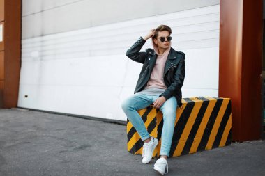 Pembe bir kazak beyaz spor ayakkabı, kot pantolon siyah ceketli trendy güneş gözlüklü genç hipster adam çizgili bir betonarme döşeme arka planı beyaz bir metal geçit üzerinde oturuyor. Amerikalı adam