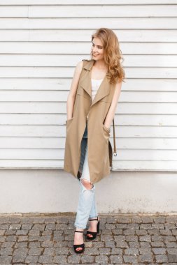 Mavi yırtık kot pantolon yüksek topuklar ve ahşap beyaz duvar poz bir deri çanta siyah ayakkabılarla şık bir T-shirt hafif yaz yeleğe sevimli neşeli genç kadın. Mutlu kız