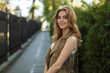 Şık yazlık elbise şık siyah deri çanta yeşil ağaçlar ve eski bir demir çit yakınında sokakta poz ile mutlu zarif genç kadın. Harika bir gülümseme ile güzel kız.