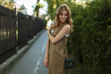 Yemyeşil ağaçlar ve bir demir vintage çit yakınında sokakta poz bir siyah deri çanta ile şık yazlık elbise sevimli çekici genç kadın. Gülümseyen güzel kız