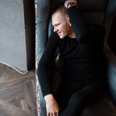 Spor ayakkabı içinde moda kot pantolon siyah şık gömlek şık mutlu genç adam odanın penceresinde yakın yumuşak bir vintage sandalyede oturur. Hafta sonu neşeli sevimli adam sahiptir.