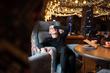 Komik neşeli genç adam şık siyah gömlekli modaya uygun kot pantolon vintage bir ahşap masa sandalye parlak ışıklar arka plan üzerinde bir kafede oturur. Hafta sonu keyfi mutlu adam.