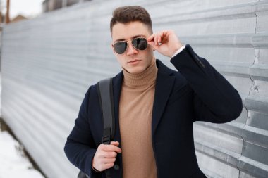 Şık yakışıklı genç hipster adam şık siyah güneş gözlüğü bej örme kazak bir sırt çantası ile şık ceket teşkil etmektedir ve metal çit yakınında sokakta kameraya benziyor. İyi adam
