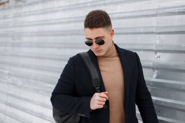 Bir sırt çantası içinde parlak metal duvarın yanında modaya uygun bir ceket bir şık saç modeli bej örme kazak içinde olan genç şık hipster adam siyah güneş gözlüğü. Bir yürüyüşe Amerikalı adam.