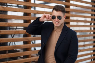 Yakışıklı genç hipster adam güzel bir gülümseme ile moda güneş gözlüğü sıcak örme kazak şık siyah paltolu ahşap duvar açık havada poz. Mutlu modern adam.