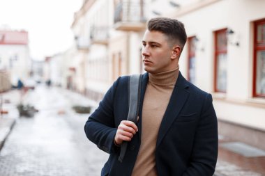 Moda yakışıklı genç bir şık siyah paltolu bir bej kazak trendy bir sırt çantası ile şık bir saç modeli ile şehrin eski binaların arka plan üzerinde pozlar ördü. İyi adam.