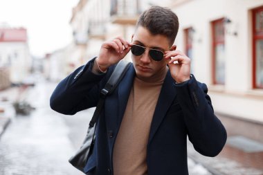 İçinde bir vintage moda güneş gözlüğü siyah paltolu şık bir saç modeli olan yakışıklı şık genç hipster adam kazak bir sırt çantası ile sıcak bir günde pozlar şehrin ördü. Kentsel Avrupa guy