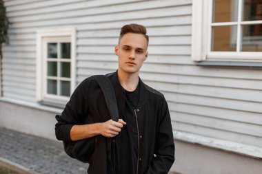 Bir T-shirt bir ahşap evin bir yaz gününde poz içinde şık bir saç modeli ile büyüleyici genç adam şık yaz ceket siyah bir sırt çantası ile. Şık adam.