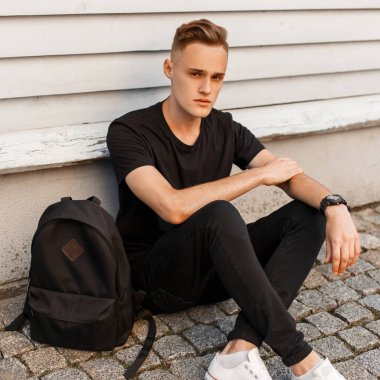 Yakışıklı genç şık siyah yaz giysileri ile şık bir sırt çantası içinde beyaz spor ayakkabı beyaz vintage bina yakınındaki oturur. Amerikan modern adam rahatlatır.