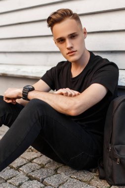 Jeans T-shirt sevimli çekici genç adam açık havada beyaz bir ahşap duvar yakınındaki bir sıcak yaz gününde oturuyor. Sevimli adam.