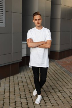 Çekici genç yakışıklı beyaz şık tişört kot ile bir saat içinde modern bir sıcak yaz gününde bina yakınındaki sokakta duruyor. Tatilde cool adam.