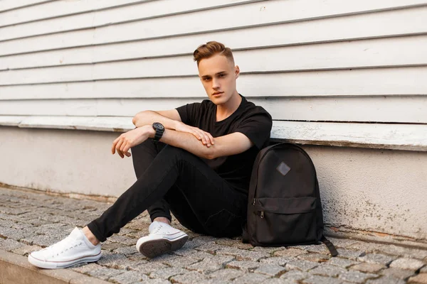 Cute young man in a black T-shirt in stylish black jeans in trendy sneakers with a black sports backpack is resting sitting on the asphalt near a white wooden vintage building. Stylish guy.