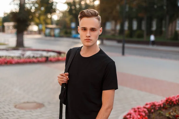 Handsome young man with a stylish hairstyle in a black fashionable t-shirt with a stylish backpack walks around the city on a background of flowers. American stylish guy.