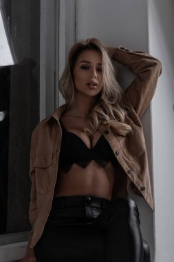 Göz alıcı güzel genç sarışın kadın kıvırcık saçlı deri şık pantolonlu bir gömlek dantel şık siyah aplik studio penceresinde yanında oturur. Şehvetli seksi kız.