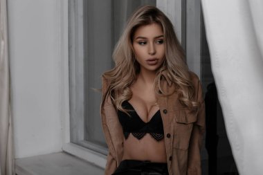 Şık dantel sütyen bir gömlek moda deri pantolonlu güzel yemyeşil memeli çok seksi genç sarışın kadınlar beyaz arka plan için pencerede yakınındaki bir odada oturur. Şehvetli şık kız.