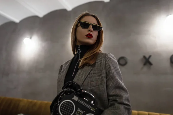 Modern hipster young woman in stylish sunglasses with a red-lips nose-ear in a checkered gray stylish jacket with a black bag in the form of a tape recorder standing indoors. Stylish future girl.