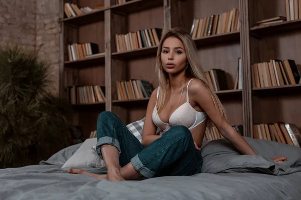 Mulher loira jovem sexy bonito em um sutiã elegante branco em jeans azul elegante sentado na cama no quarto. Modelo menina bonita está descansando dentro de casa perto da parede do vintage com livros . — Fotografia de Stock