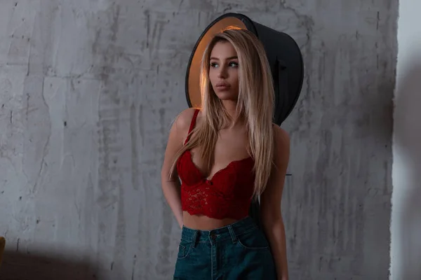 Young sexy pretty blonde in trendy fashion vintage blue jeans in a luxurious red lace bra is standing indoors near a metal light lamp. Sensual girl with a beautiful slim body. — Stock Photo, Image