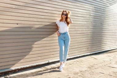 Güzel güzel hipster genç kadın güneş gözlüğü vintage kot pantolon beyaz spor ayakkabı şık beyaz t-shirt ahşap duvara yakın duruyor. Modern Amerikalı batımında.