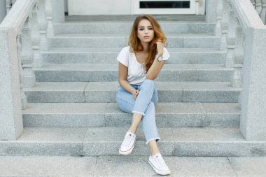 Bir beyaz genç Amerikalı kadın vintage bir bina basamaklarında oturan beyaz trendy spor ayakkabı jeans T-shirt rahatlatır. Sevimli Avrupa kız modeli diğer sahiptir. Stil kadın giyim bahar.