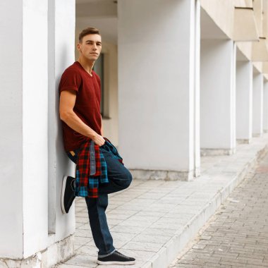 Genç şık şık kırmızı t-shirt açık havada bir yaz gününde bir beyaz vintage bina yakın poz spor ayakkabı moda mavi kot pantolon içinde trendy ekose gömlekli adam. Yakışıklı adam bir yürüyüş sahiptir.