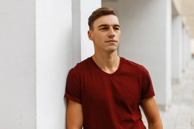 Neşeli genç yakışıklı bir trendy kırmızı T-shirt şık bir saç modeli ile modern beyaz bina yakınında sokakta duruyor. Dinlenme ve bir sıcak yaz günü zevk şirin neşeli adam.