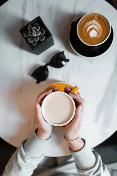 Vrouwelijke handen met een beker met warme koffie in het café. Jonge vrouw zit in de ochtend binnen. Bovenaanzicht van de tabel met koffie en zonnebril. — Stockfoto