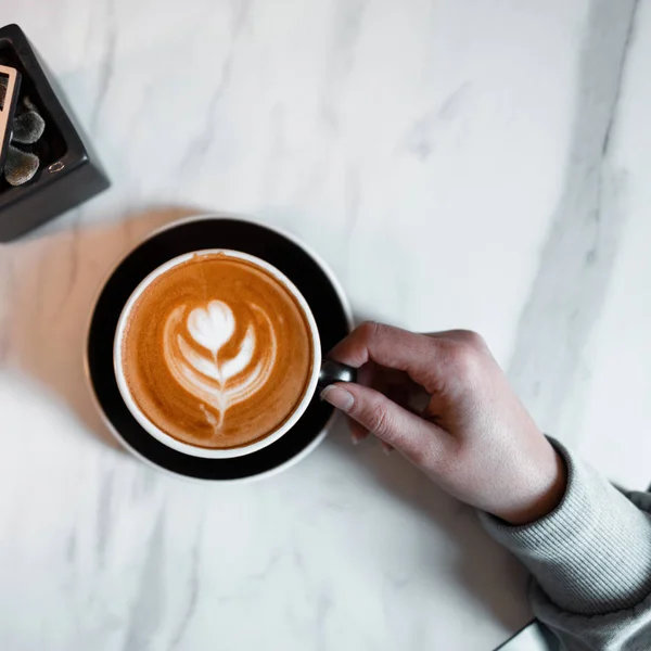 Close-up van vrouwelijke hand hebt een kopje cappuccino op een vintage houten tafelblad in een koffieshop. Ochtend koffiepauze. Van bovenaf bekijken. — Stockfoto