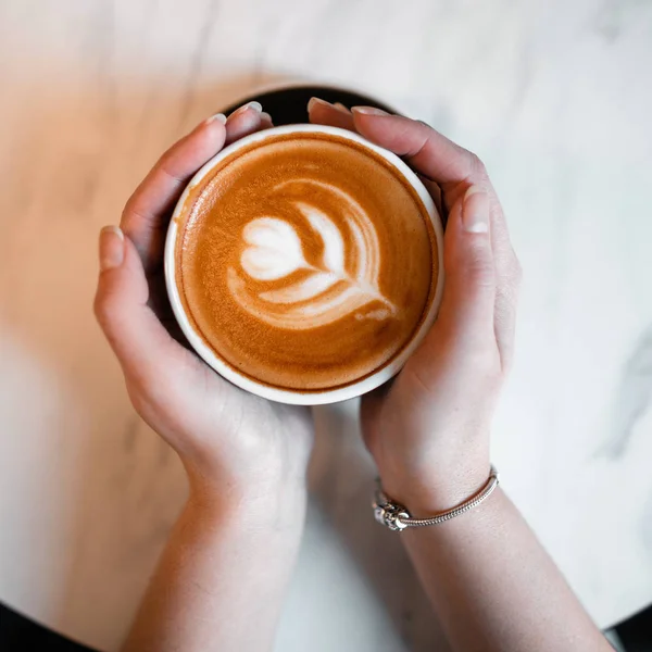 Vrouwelijke handen met een kop met een heerlijke latte op de achtergrond van een vintage tafel. Ochtendpauze koffie in een café. Close-up. Van bovenaf bekijken. — Stockfoto