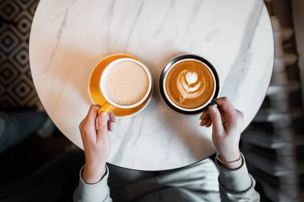 Vrouw houdt twee kopjes van warme dranken. Warme smakelijke latte en cappuccino in vrouwelijke handen. Lunchpauze voor koffie. Van bovenaf bekijken. — Stockfoto