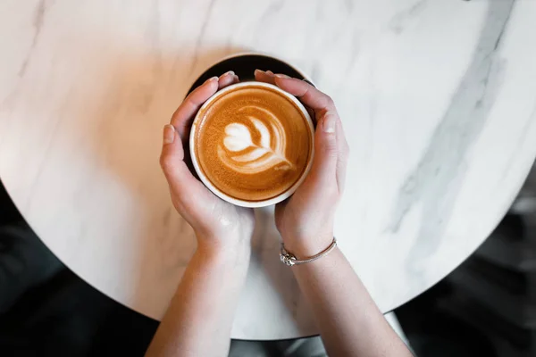 Vrouwelijke handen met een kop warme koffie met schuim over een houten tafel. Bekijk van bovenaf. Geweldige ochtend. — Stockfoto