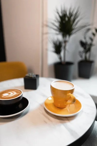 Na stole v krásném retro kavárně je černý hrnek horké latte a cappuccino žlutý hrnek. Ranní kávová přestávka v gourmet café. Odpočinek s přáteli v kavárně. — Stock fotografie