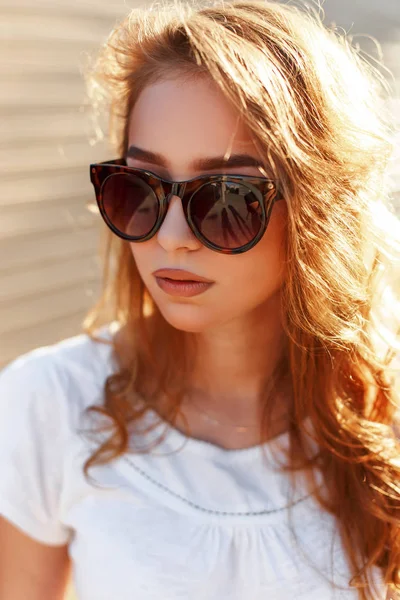 Retrato de mujer joven hipster bastante lindo en gafas de sol de moda en blanco verano elegante camiseta con pelo rubio contra el fondo de una pared de madera. Hermosa chica en un paseo disfruta del sol . —  Fotos de Stock