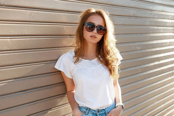 Young modern hipster woman in stylish sunglasses in a summer fashion white t-shirt in trendy jeans is standing near a wooden vintage wall on a sunny spring day. Cute girl resting outdoors. — Stock Photo, Image